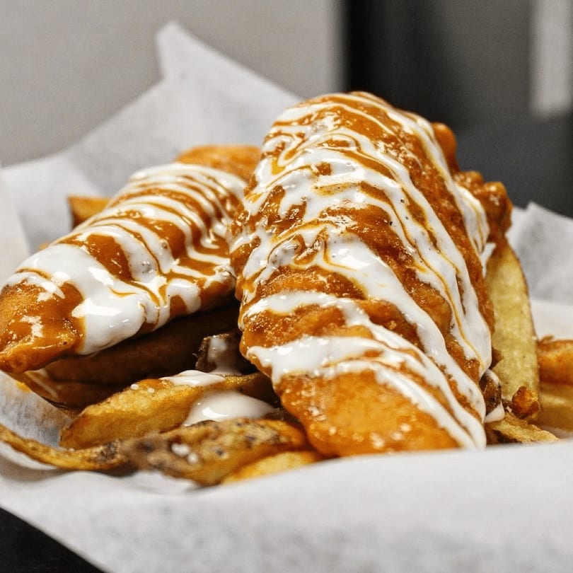 Two golden-fried cheesesteak rolls drizzled with white and orange sauces, served with fries.