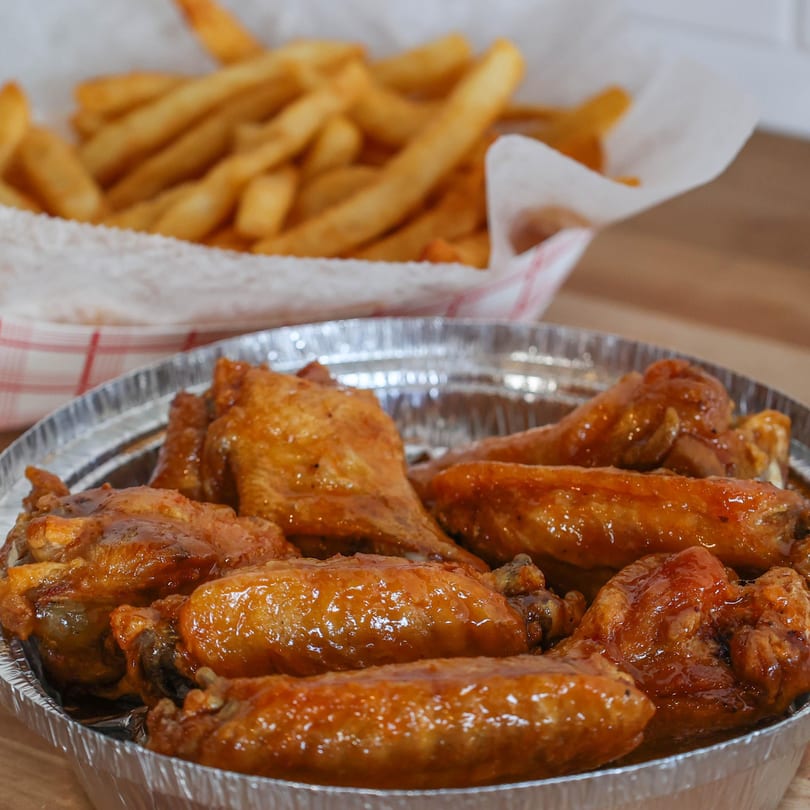 A foil container of glazed chicken wings with a basket of french fries in the background.