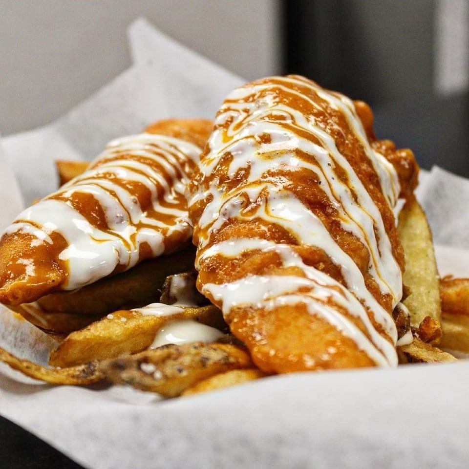 Two Wiz Kidz Cheesesteaks, drizzled with white and orange sauces, served with fries in a paper-lined basket.