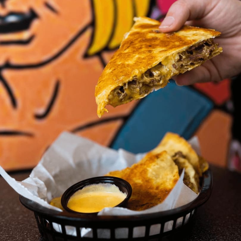 Hand holding a cheesy cheesesteak quesadilla slice, with more in a basket and cheese sauce.